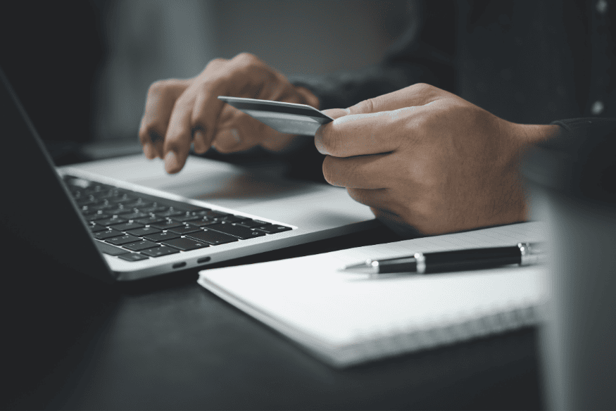 Person typing on a laptop while holding a credit card, next to a notebook and pen