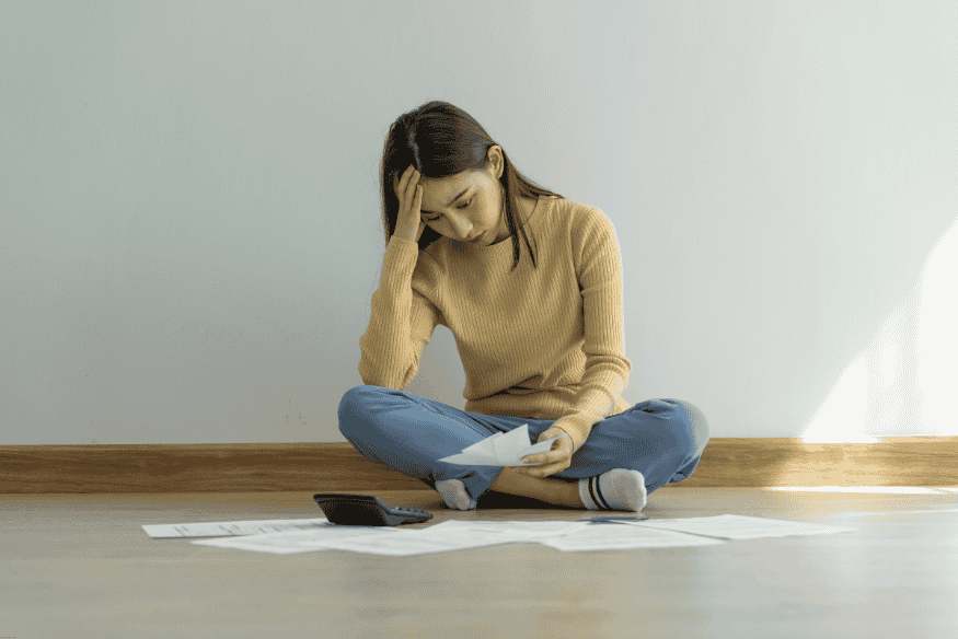 Woman sitting on the floor looking stressed while reviewing bills and using a calculator
