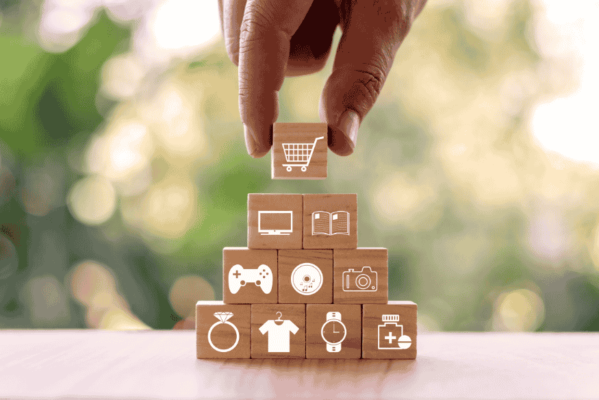 Hand placing a wooden block with a shopping cart icon on top of blocks with icons representing consumer goods