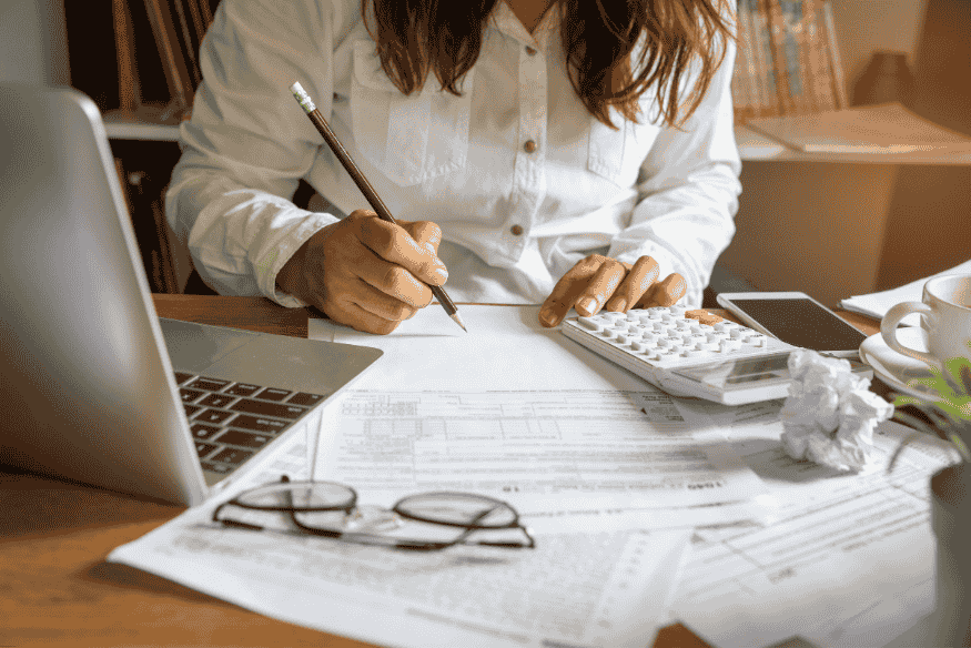Woman using a calculator and taking financial notes at a desk with documents and a laptop